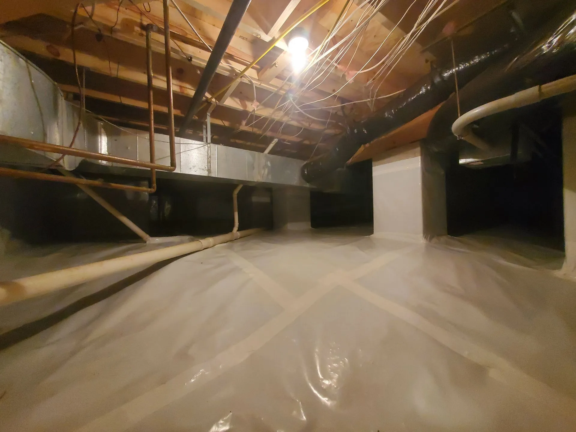 A crawl space with a plastic vapor barrier on the floor, exposed pipes, ducts, and wiring, illuminated by a single overhead light.