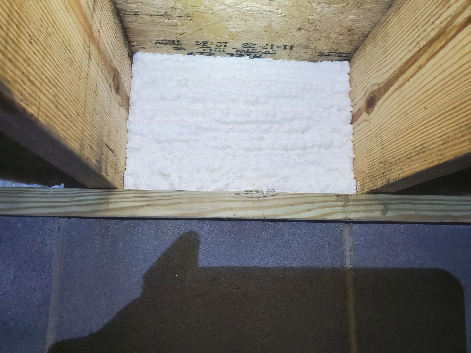 Spray foam insulation applied between wooden joists in a floor or ceiling cavity, with a shadow cast on the lower portion of the area.