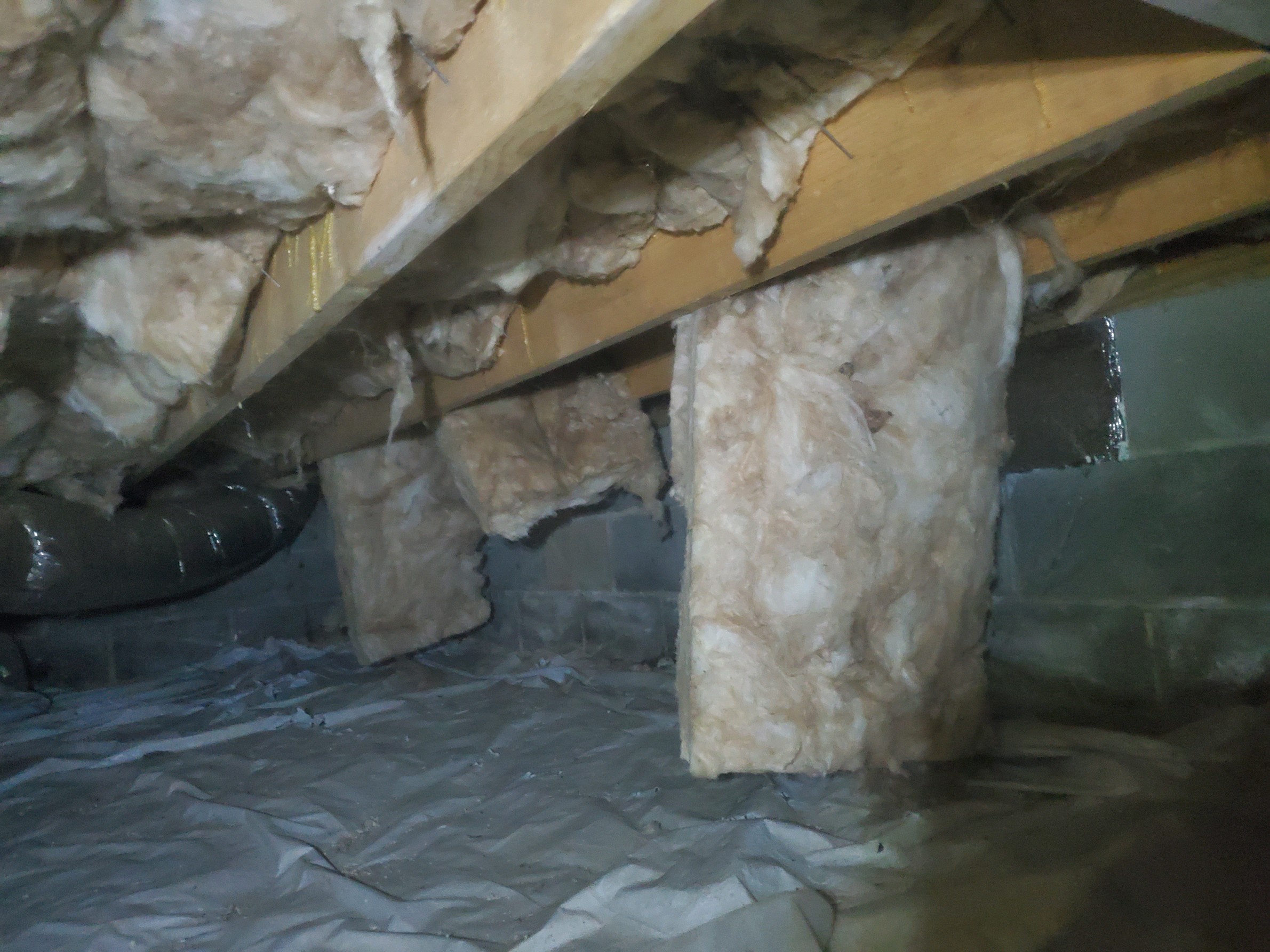 Fiberglass insulation hangs loosely from floor joists and supports in a crawl space, with a plastic vapor barrier covering the ground and some cinder block walls visible.