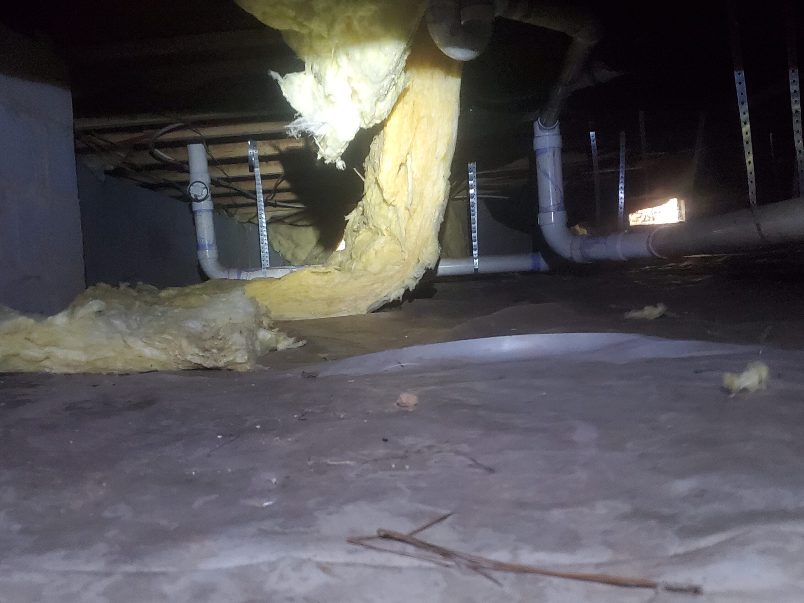 Crawl space with exposed pipes and yellow insulation hanging down, illuminated by a flashlight beam.