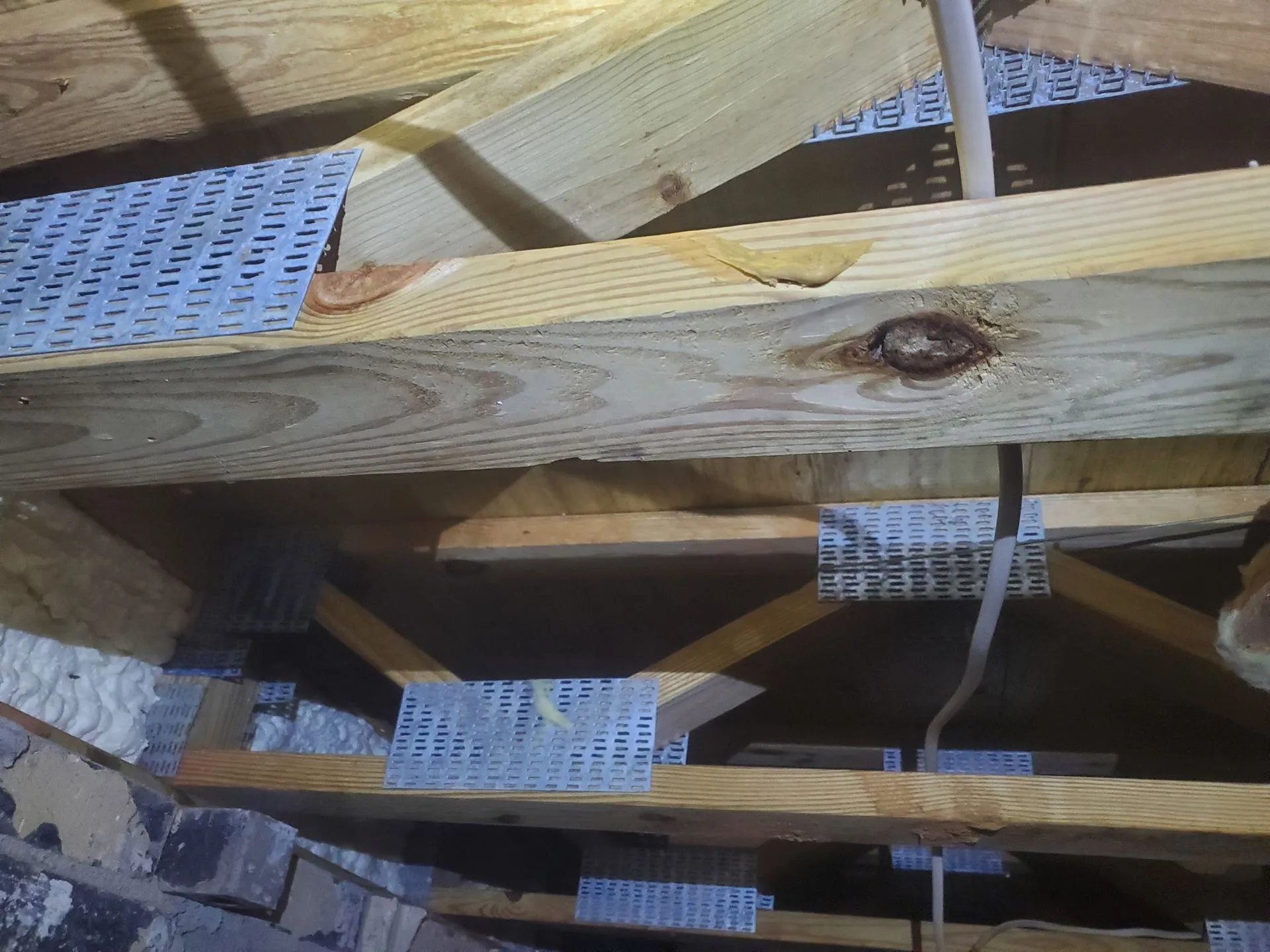 Wooden beams with metal plates provide structural reinforcement in a ceiling or attic space, where insulation is visible on the left and wiring runs across the beams, creating an environment that may benefit from mold removal.